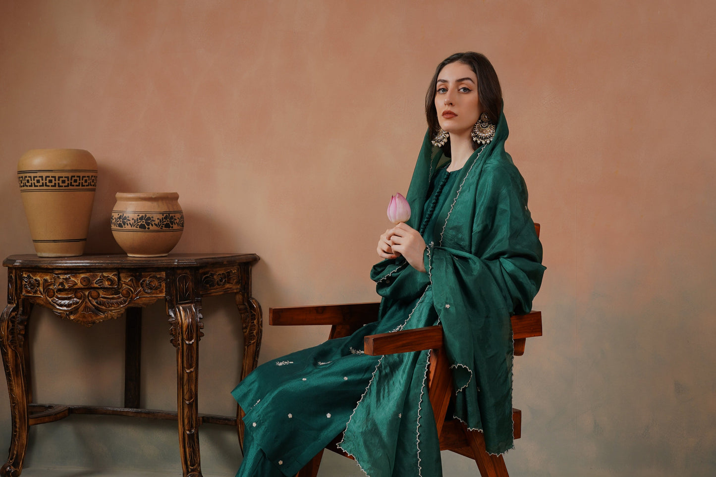 Woman in a green outfit sitting on a wooden chair against a pinkish-beige wall.