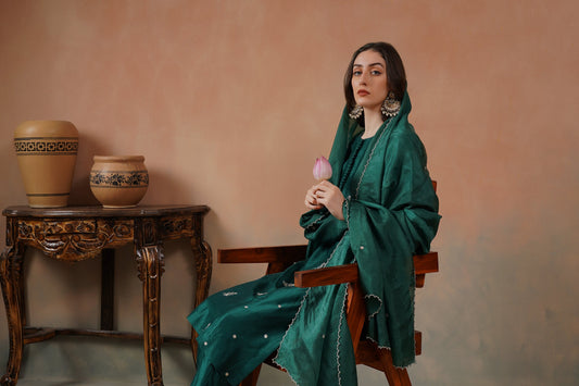 Woman in a green outfit sitting on a wooden chair against a pinkish-beige wall.